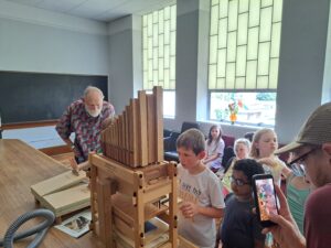 The older organist pumps the Orgelkit instrument while a few children stand by to watch and listen.